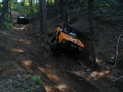 1988 Ford Bronco - Two off-road vehicles navigate a challenging, wooded trail with steep inclines and loose dirt.