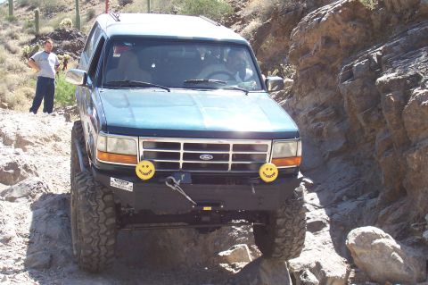 1992 Ford Bronco - A blue 1992 Ford Bronco is shown off-roading through a narrow, rocky canyon with a person standing on the side.