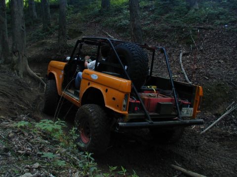 1988 Ford Bronco - An orange SUV navigates a wooded trail, equipped with a roll cage and external fuel cans.