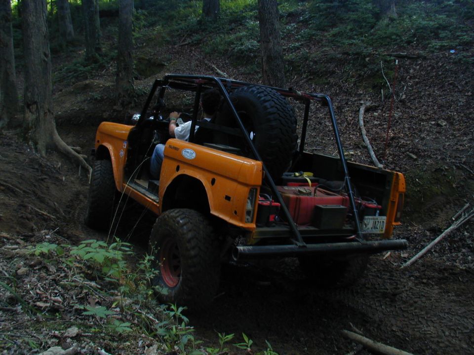 Orange SUV with a roll cage driving on a dirt trail in the woods. Spare tire mounted in the back
