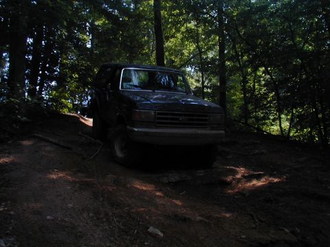 1988 Ford Bronco - A Ford Bronco navigates a wooded trail, its front end angled towards the camera on the uneven terrain.
