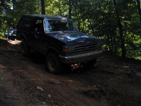 1988 Ford Bronco - A dark-colored Ford Bronco navigates an off-road trail through a wooded area.
