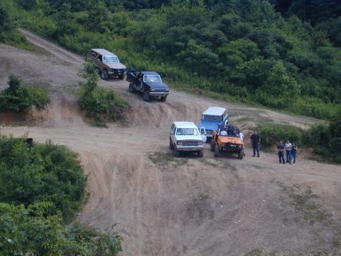 1988 Ford Bronco - Several off-road vehicles navigate a dirt track surrounded by lush green trees.