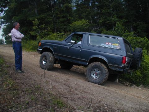 1988 Ford Bronco - A man stands by a dark blue 1988 Ford Bronco as it navigates a dirt road, with a BFGoodrich sticker visible on the rear window.