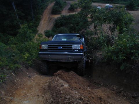 1988 Ford Bronco - A dark-colored Ford truck navigates a steep, muddy off-road trail with trees and foliage surrounding it.