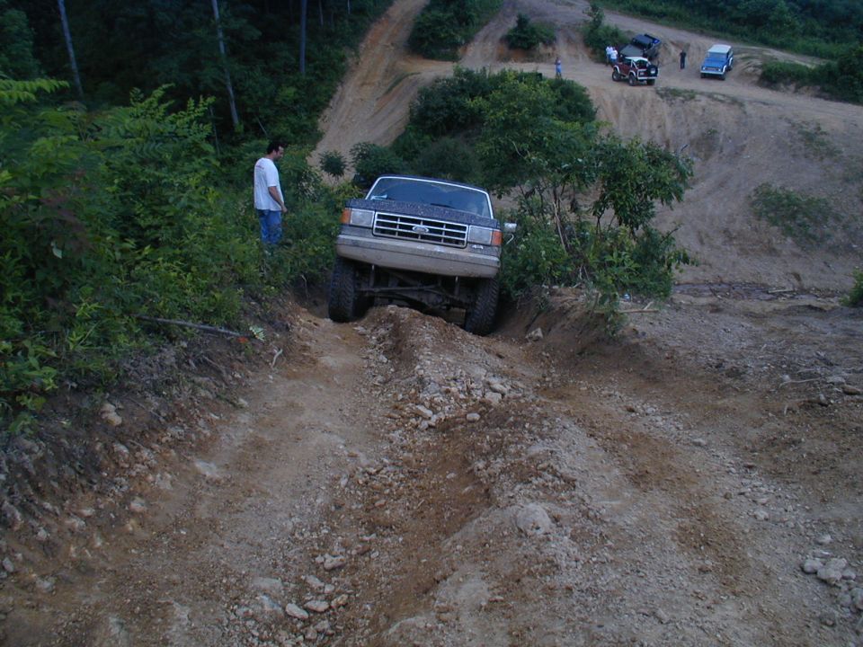 Ford Bronco climbing a dirt hill off-road, with a person watching. Other vehicles are visible in the background