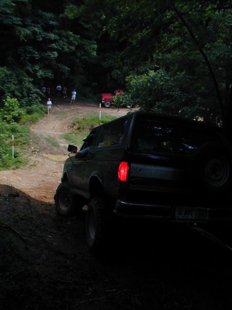 1988 Ford Bronco - A dark SUV navigates a muddy, wooded trail with other vehicles and people in the background.