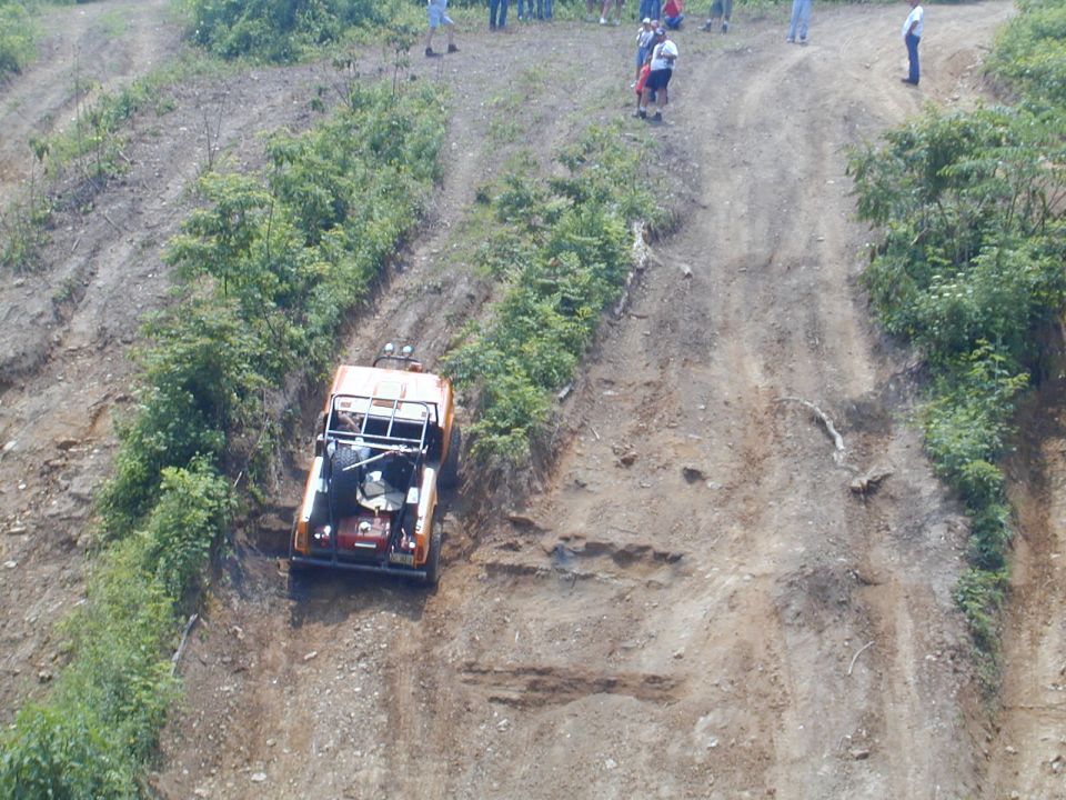 Orange off-road vehicle with a roll cage and spare tire on the back is climbing a steep, muddy hill. Spectators watch from the top