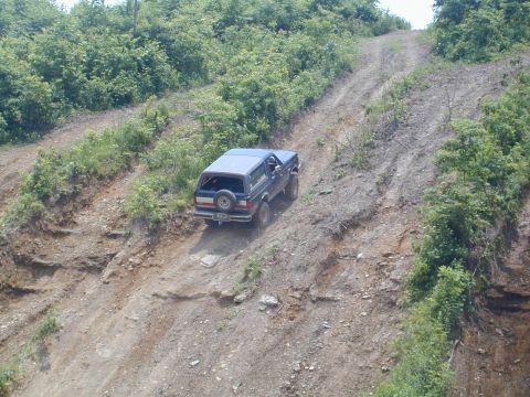 1988 Ford Bronco - A dark blue two-door SUV with a rear-mounted spare tire descends a steep, deeply rutted dirt trail cut into an eroded hillside surrounded b…