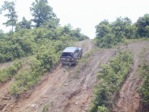 1988 Ford Bronco - A dark 1988 Ford Bronco navigates an off-road trail, its rear profile visible as it ascends a steep dirt incline.