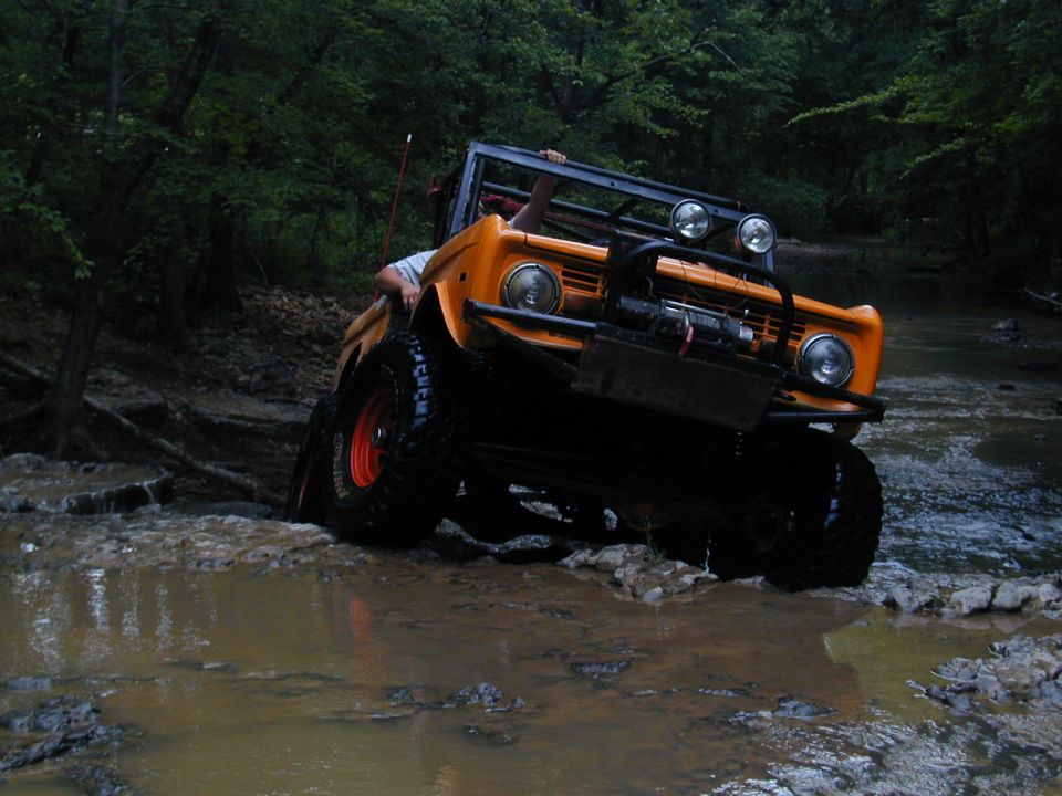 Orange Bronco driving through mud and water on a trail. It has a winch and extra lights on the front