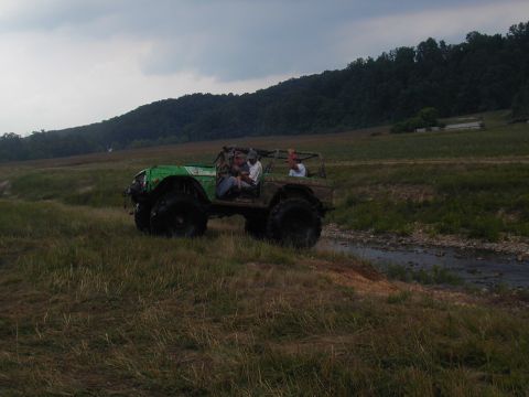 1988 Ford Bronco - An off-road vehicle with people inside navigates a muddy, grassy terrain under a cloudy sky.
