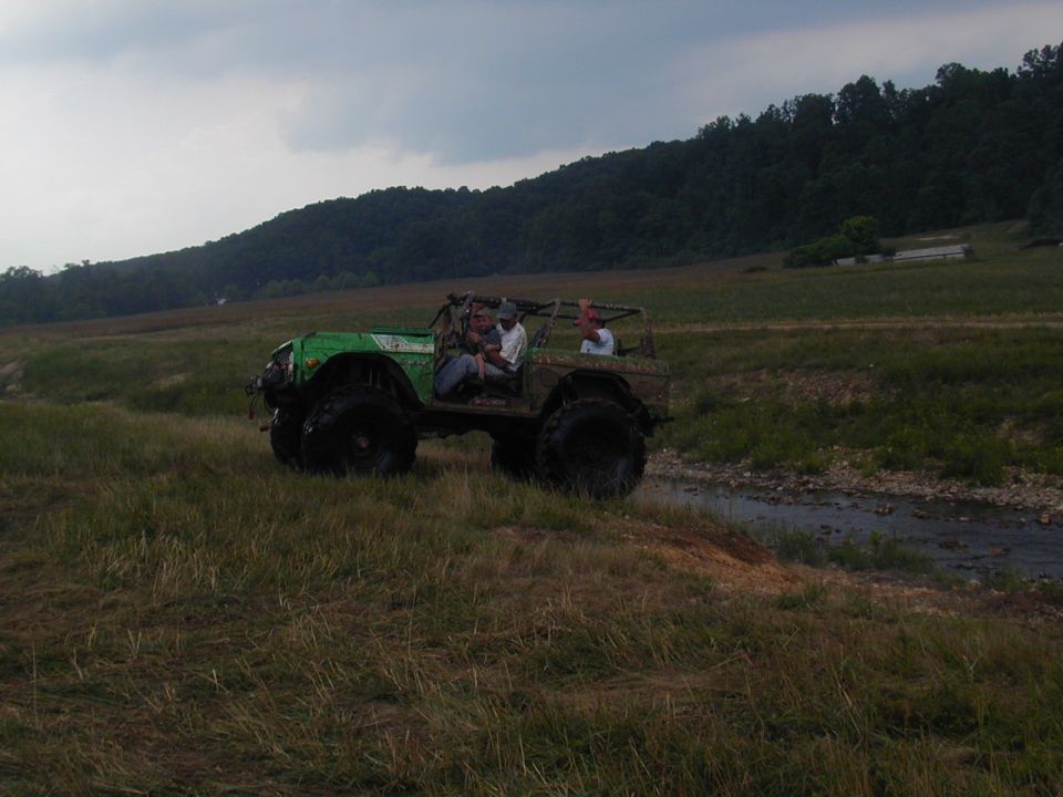 Green off-road vehicle with large tires driving through a grassy field near a stream