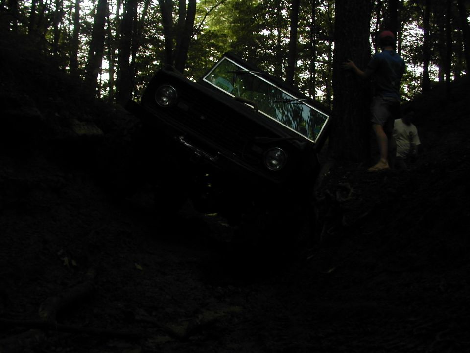 Black SUV tilted on a muddy hill in woods, two people nearby