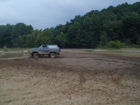 1988 Ford Bronco - A blue Ford Bronco navigates a muddy terrain with trees in the background.