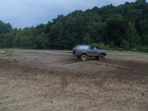 1988 Ford Bronco - A dark blue Ford Bronco navigates a muddy terrain with a wooded area behind it.