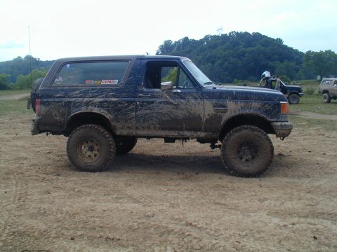 1988 Ford Bronco - A dark blue two-door Ford Bronco SUV shown in side profile, heavily splattered with mud and lifted on large off-road tires with black wheel…