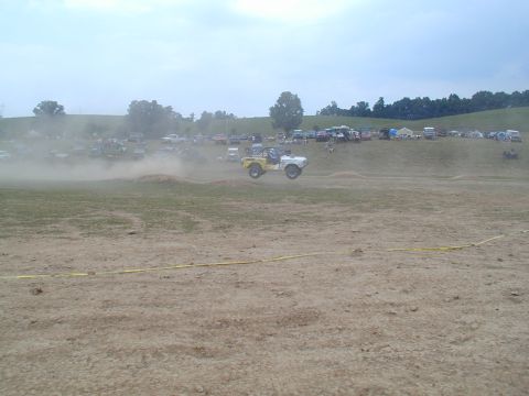 1988 Ford Bronco - A 1988 Ford Bronco takes flight over a dirt mound at an off-road event, with spectators and other vehicles in the background.