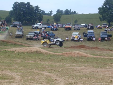 1988 Ford Bronco - An off-road event shows multiple vehicles gathered, with one jeep performing a jump over a dirt ramp.