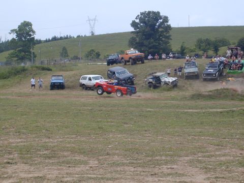 1988 Ford Bronco - A red off-road vehicle soars through the air during an event, with multiple other vehicles and spectators visible in the background.