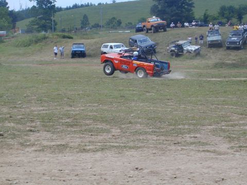 1988 Ford Bronco - Multiple vehicles compete in an off-road event on a grassy course with spectators watching from the hillside.