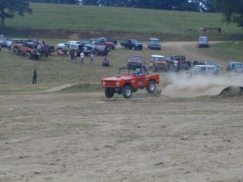1988 Ford Bronco - An orange Ford Bronco jumps over a dirt mound during an off-road event, with other vehicles and spectators in the background.
