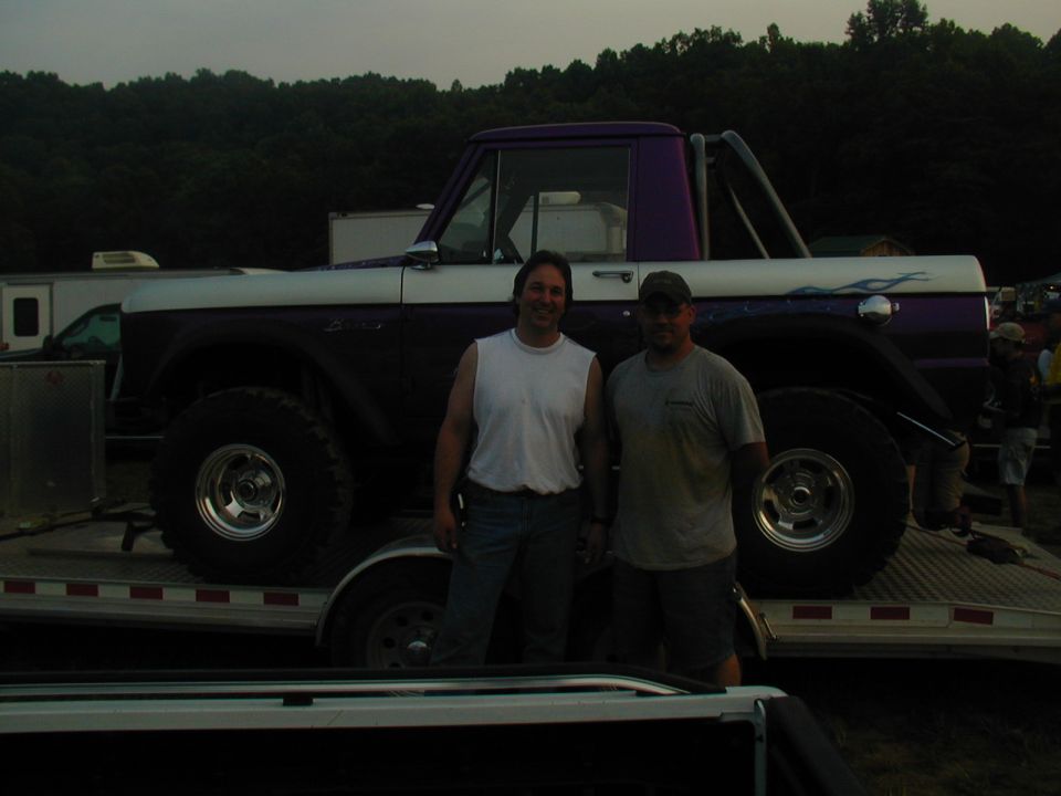 Two guys stand in front of a lifted Ford Bronco on a trailer. It has a purple and white paint job with flames on the side