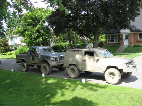 1986 Ford Bronco - Two modified SUVs, one dark green and one muddy tan, are parked on a residential street.