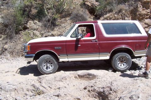 1992 Ford Bronco - A maroon and white 1992 Ford Bronco navigates a , rocky path in a desert landscape.