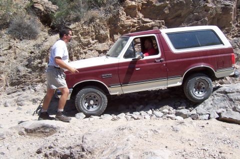 1992 Ford Bronco - Man pushes a 1992 Ford Bronco through a rocky off-road trail in a desert landscape.