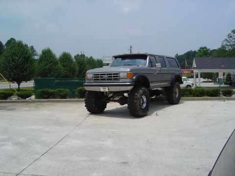 1986 Ford Bronco - This lifted Ford Bronco is parked in a lot with trees and a building in the background.