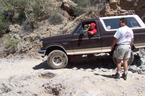 1992 Ford Bronco - Side view of a dark brown 1990s full-size Ford Bronco with a white hardtop and tan lower stripe working up a loose rock ledge on a narrow d…
