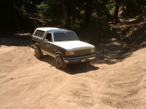 1990 Ford Bronco - A two-tone Ford Bronco with an aftermarket grille drives on a dirt path surrounded by trees.