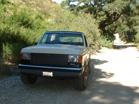 1990 Ford Bronco - A tan 1990 Ford Bronco 4x4 is parked on a gravelly dirt road surrounded by trees and brush.