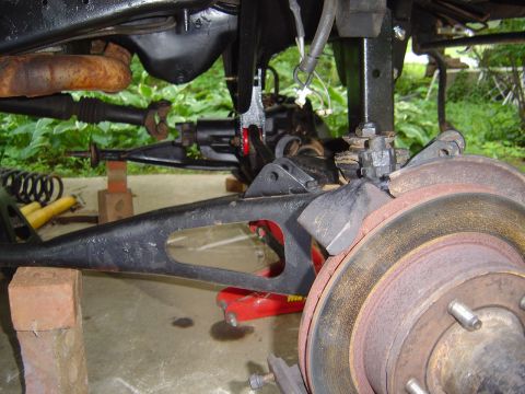 1986 Ford Bronco - Close-up underside view of a vehicle's front suspension and brake components, showing a rusty brake rotor with wheel studs, a black cast co…
