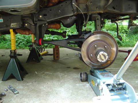 1986 Ford Bronco - Underside view of a vehicle raised on green jack stands and a gray hydraulic floor jack, showing the rear brake drum/hub, leaf spring, cont…