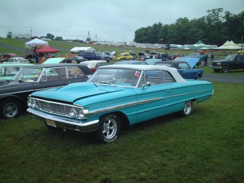 1967 Ford Galaxie - A turquoise 1964 Ford Galaxie convertible with a hood scoop is parked on the grass at an outdoor car show.