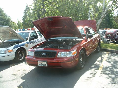 1999 Ford Crown Victoria - A red 1999 Ford Crown Victoria is displayed with its hood open, parked next to a police car and a vintage vehicle.