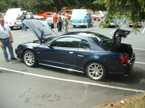 1999 Ford Crown Victoria - A dark blue Ford Mustang GT coupe is displayed with its hood and trunk open at an outdoor car show.