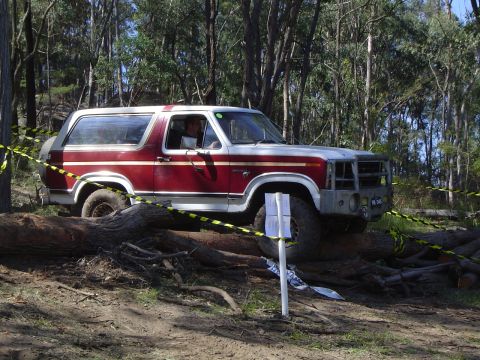 1982 Ford Bronco - A red and white Ford Bronco navigates an off-road course, climbing over fallen trees.