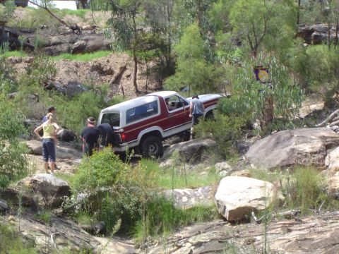 1982 Ford Bronco - A red and white Ford Bronco is shown off-roading on a challenging rocky path with several people observing.