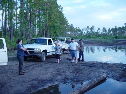 1989 Ford Bronco - People gather around trucks in a muddy off-road area with trees and water.