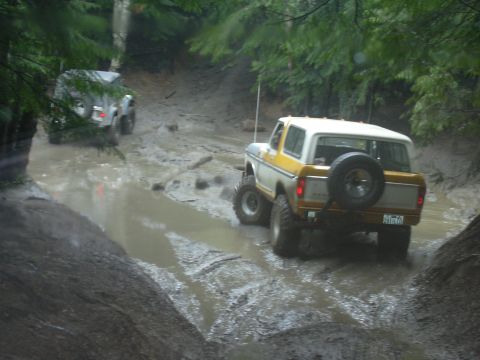 1991 Ford Bronco - Two off-road vehicles navigate a challenging muddy path in a wooded area.