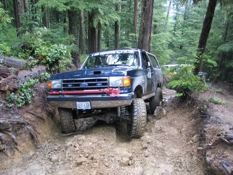 1991 Ford Bronco - A dark blue 1991 Ford Bronco navigates a challenging muddy off-road trail through a dense forest.