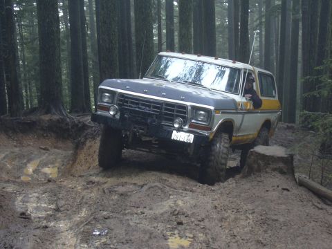 1991 Ford Bronco - Ford Bronco navigates a muddy path in a dense forest, with a tree stump nearby.