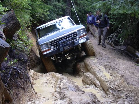 1991 Ford Bronco - A gray Ford Bronco tackles a challenging muddy off-road course, with onlookers observing the difficult terrain.