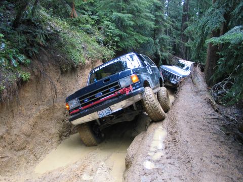 1991 Ford Bronco - Two Ford Broncos tackle a challenging muddy off-road trail in a dense forest.