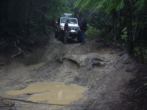 1991 Ford Bronco - A white Jeep Wrangler navigates a challenging muddy trail in a wooded area.