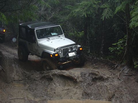 1991 Ford Bronco - A white Jeep Wrangler TJ tackles a challenging muddy off-road course, with another vehicle visible in the background.