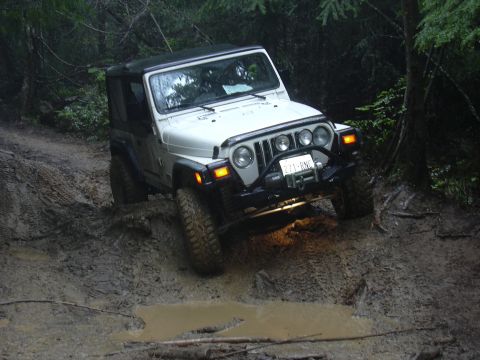 1991 Ford Bronco - White Jeep Wrangler TJ tackles a challenging muddy trail in a wooded area.